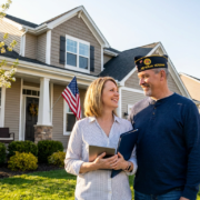 smiling couple, a veteran, and real estate agent discussing va home loans may, in front of a new house.