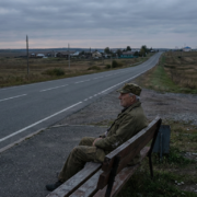 elderly veteran in fatigue uniform sitting on a bench beside a long road, highlighting veteran transportation barriers.