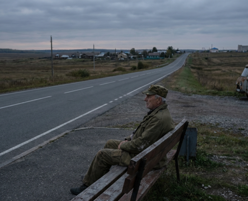 elderly veteran in fatigue uniform sitting on a bench beside a long road, highlighting veteran transportation barriers.