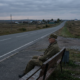elderly veteran in fatigue uniform sitting on a bench beside a long road, highlighting veteran transportation barriers.