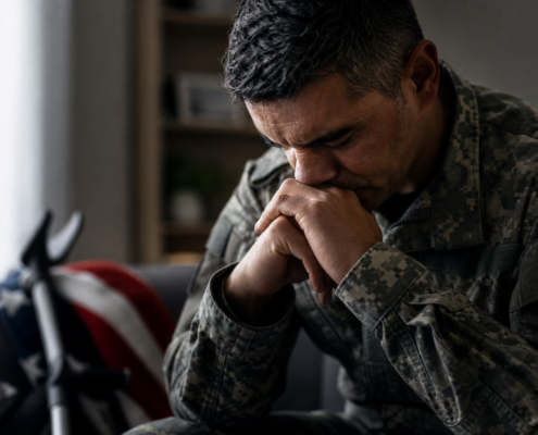 distressed veteran in uniform, hands clasped, indicating at-risk veterans warning signs; flag and crutches behind him.