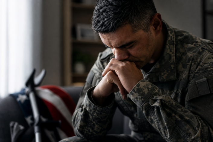 distressed veteran in uniform, hands clasped, indicating at-risk veterans warning signs; flag and crutches behind him.