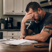 stressed veteran with american flag patch on shirt, head in hand, surrounded by papers, showing financial hardship signs.