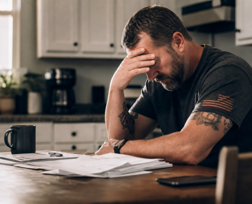 stressed veteran with american flag patch on shirt, head in hand, surrounded by papers, showing financial hardship signs.