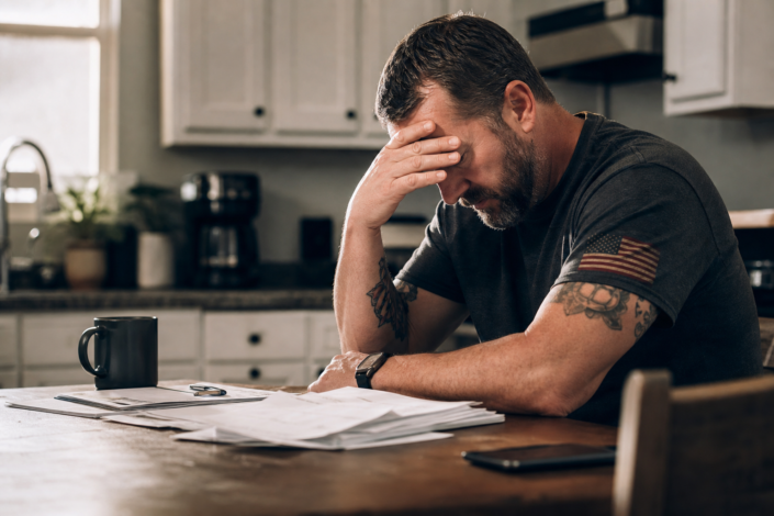stressed veteran with american flag patch on shirt, head in hand, surrounded by papers, showing financial hardship signs.