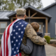 military member with flag embracing child, looking at a modern house for temporary housing for veterans.